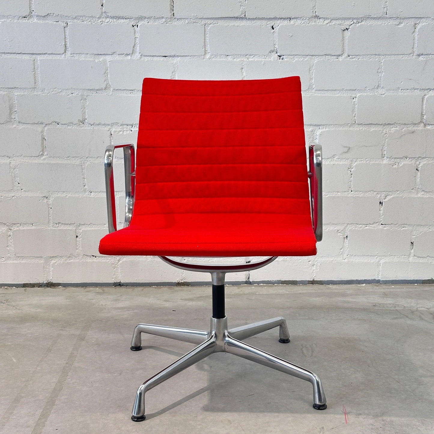 Vitra EA 108 Drehstuhl mit rotem Hopsak-Bezug und poliertem Aluminiumgestell vor weißer Ziegelwand