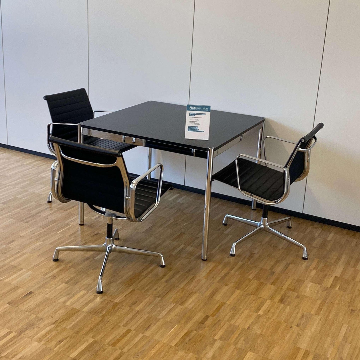 USM Haller Tisch 100x100cm with black oak top and chrome frame surrounded by three modern black office chairs on wooden floor