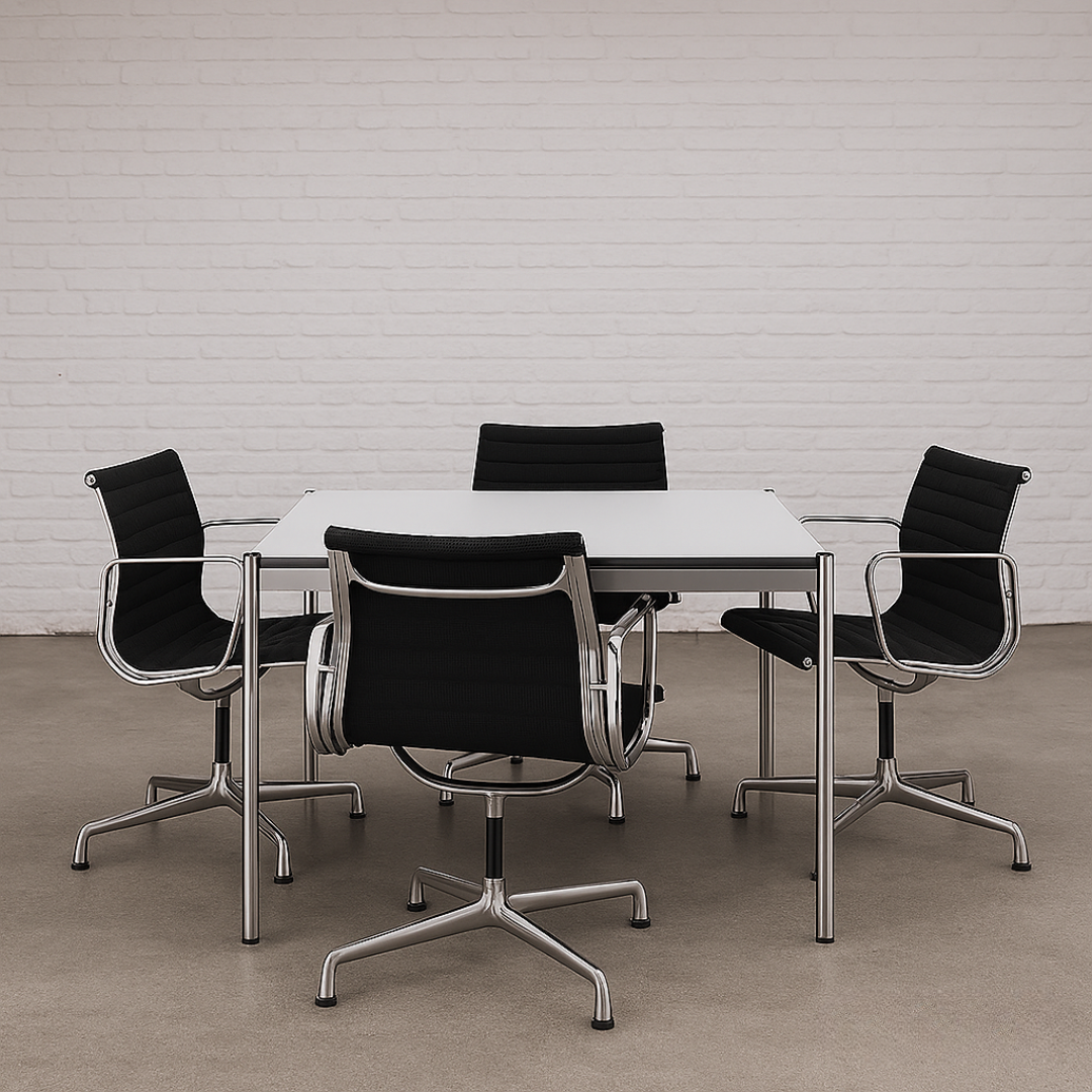 USM Haller Tisch 100x100cm perlgrau mit Chromgestell und schwarzem Bürostuhl-Set vor weißer Ziegelwand
