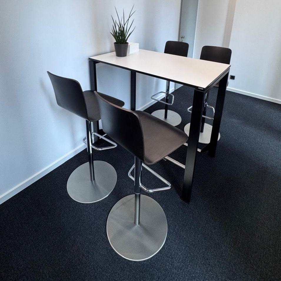 Vario Icon Bistrotisch with premium white tabletop, black frame, stainless steel footrest, and four black bar stools in modern office setting