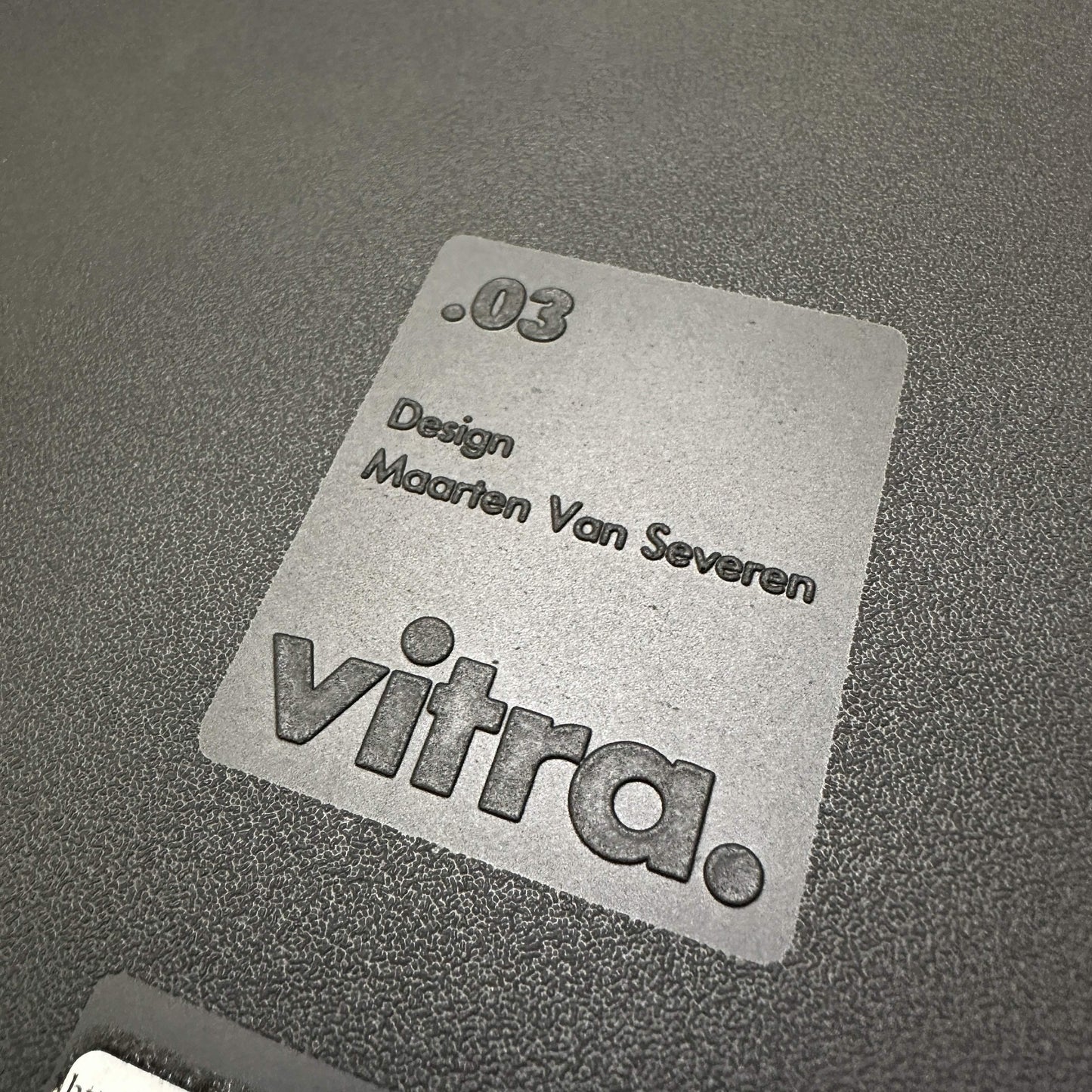 Close-up of the Vitra .03 Besucherstuhl designer label on matte black plastic seat.