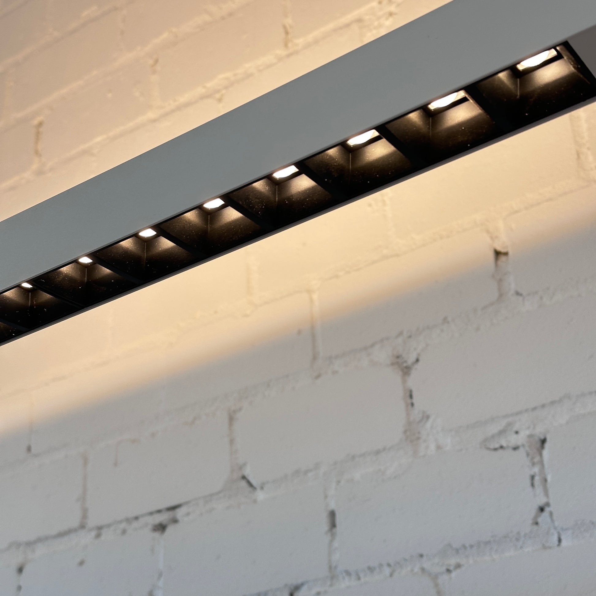 Weiße Artemide Econ Office Floor L LED-Leuchte aus Aluminium mit warmweißem Licht an einer Ziegelwand.