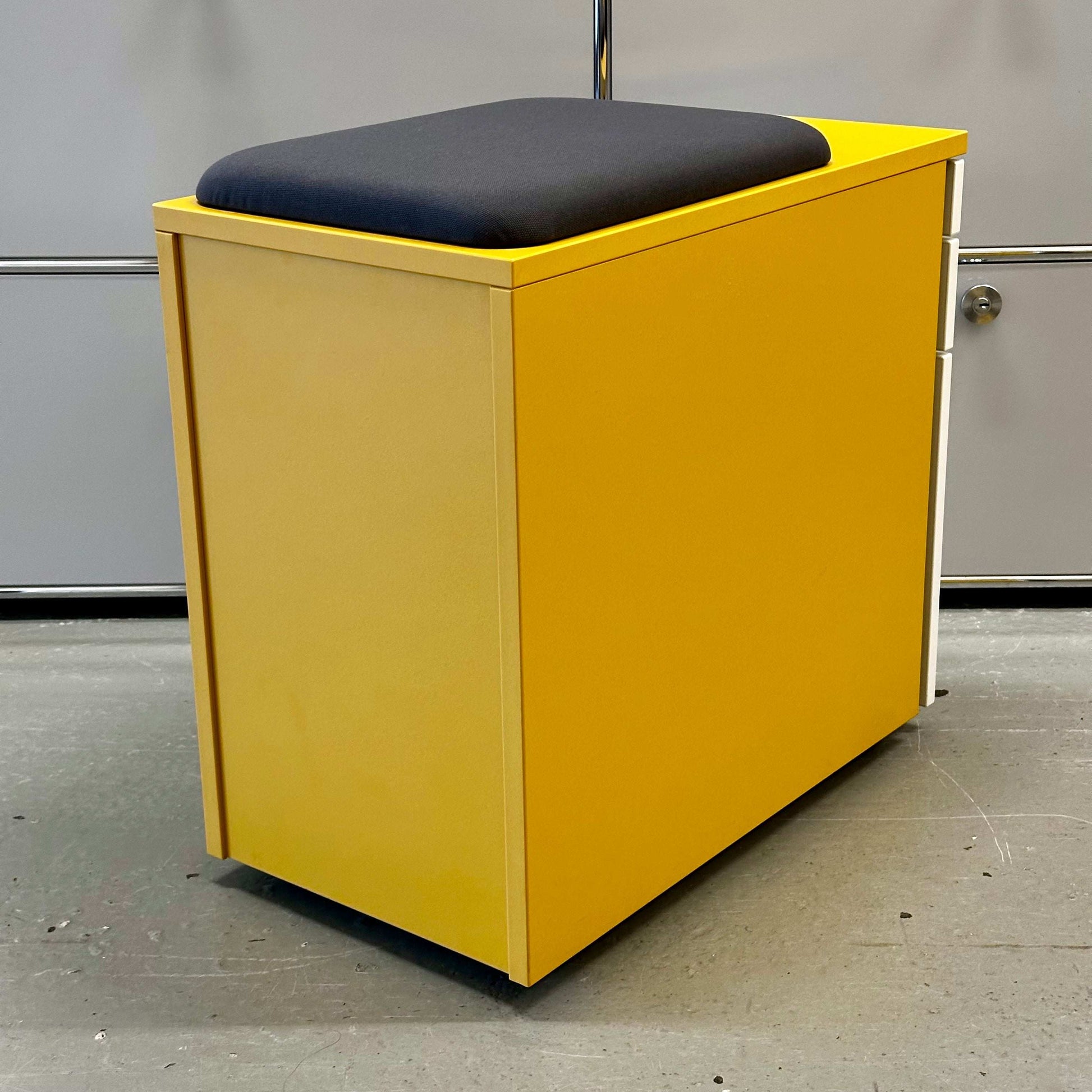 Rollcontainer CT in mustard yellow melamine with white front, three drawers, anthracite fabric seat cushion, vertical handle, locking system, and hard castors.