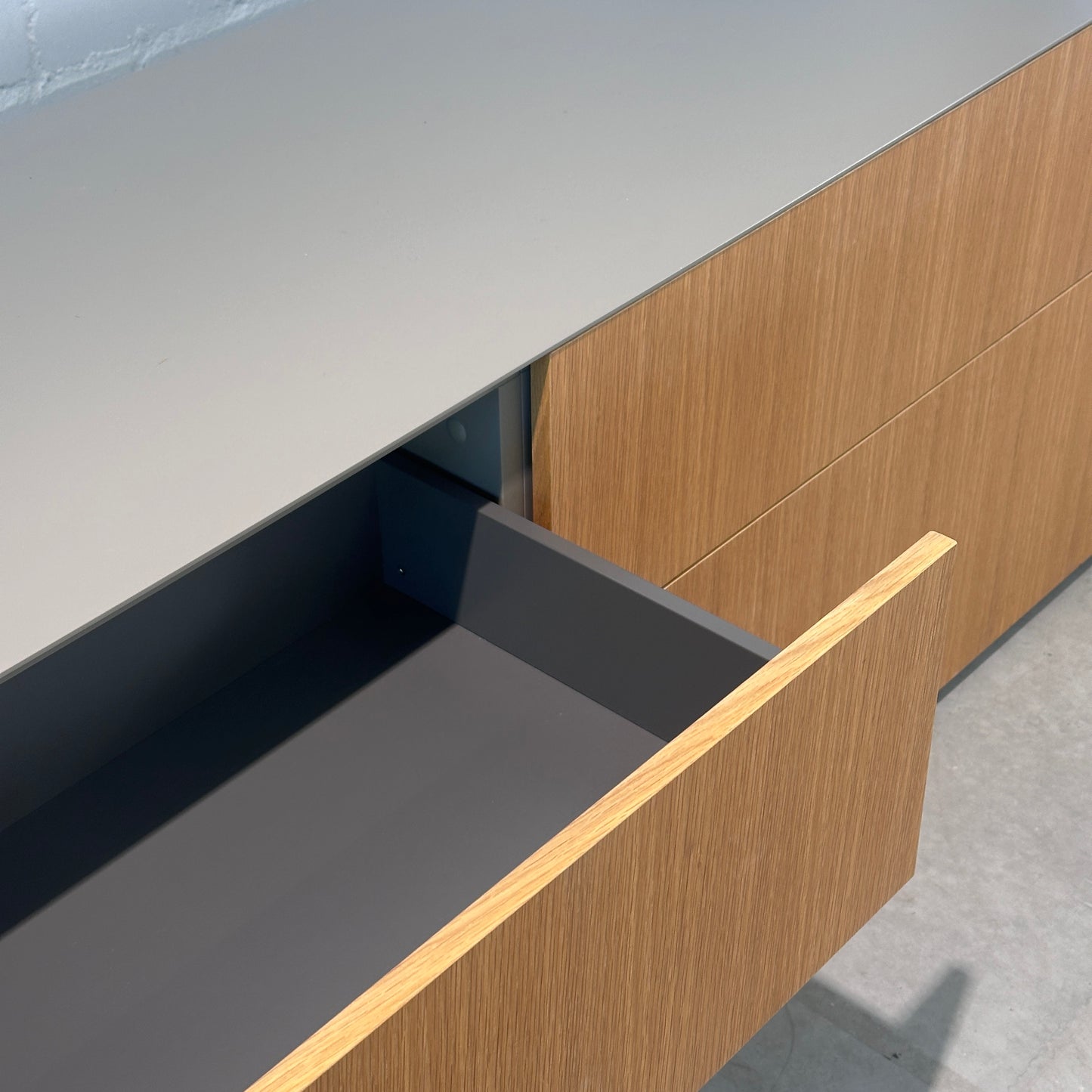 Blum Sideboard mit warmgrauer Oberfläche und Eichenfurnier-Fronten, eine Schublade offen.