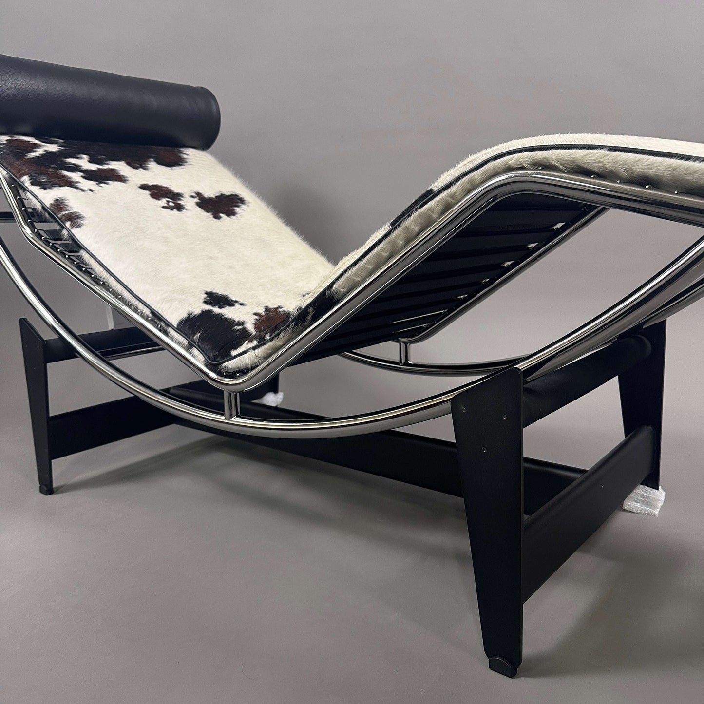 Cassina LC4 Chaiselongue Liege mit schwarz-weiß geflecktem Kuhfell und schwarzer Leder-Nackenrolle auf verstellbarem Metallrahmen und schwarzem Holzgestell