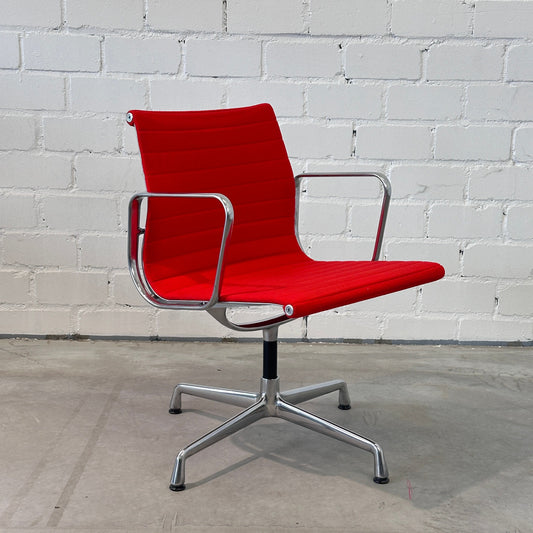 Vitra EA 108 Drehstuhl mit rotem Hopsak-Bezug und poliertem Aluminiumgestell vor weißer Ziegelwand