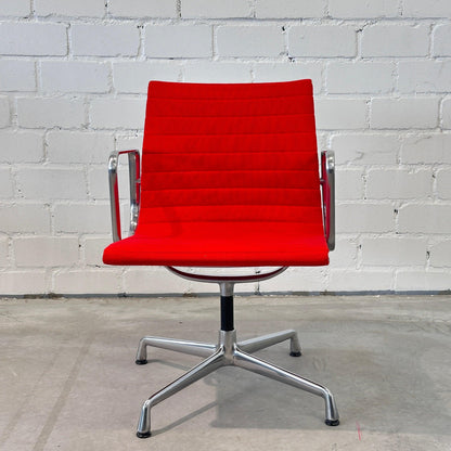 Vitra EA 108 Drehstuhl mit rotem Hopsak-Bezug und poliertem Aluminiumgestell vor weißer Ziegelwand