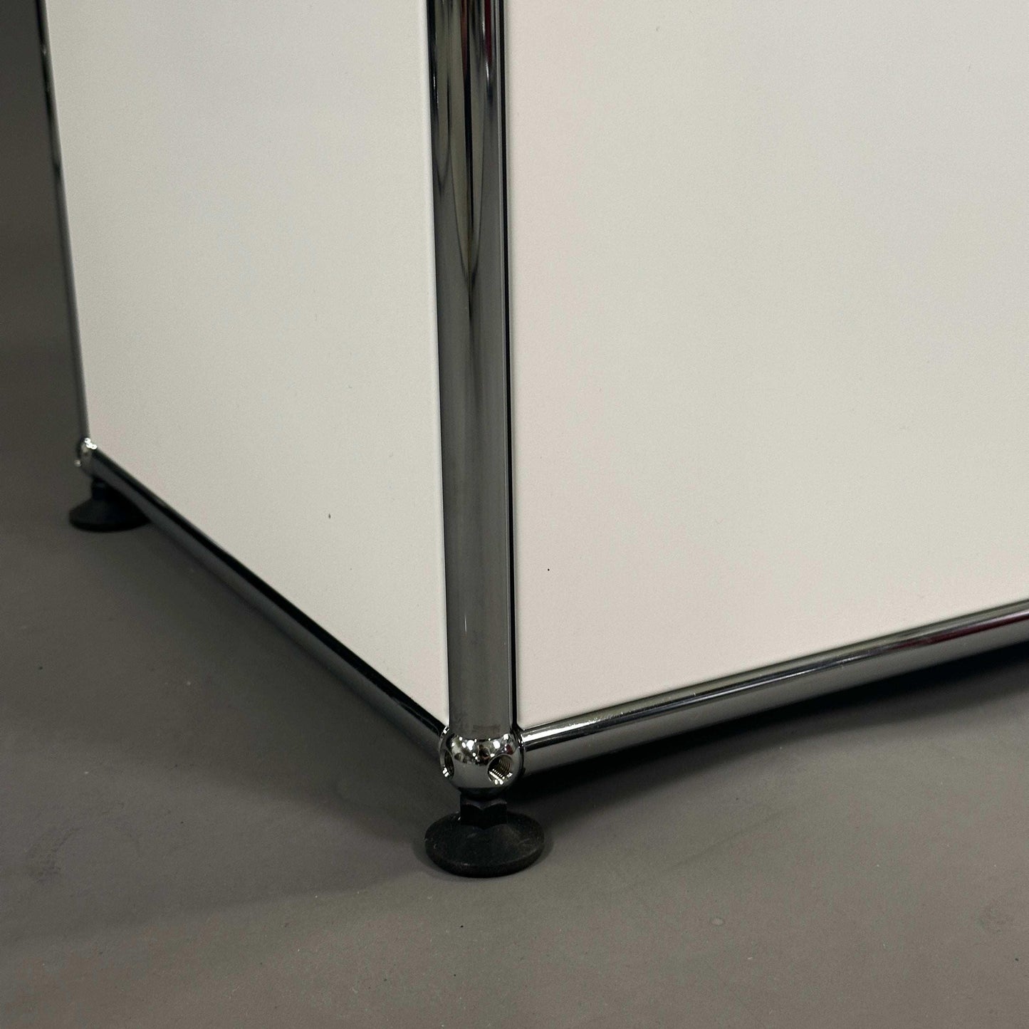 Detailaufnahme der unteren Ecke eines weißen USM Haller Highboards mit Chrom-Stahlrahmen und verstellbarem Fuß
