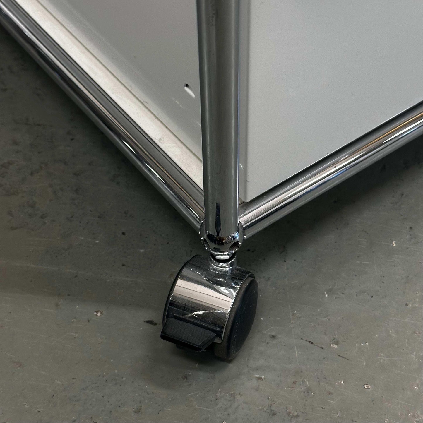 Close-up of white USM Haller Rollcontainer with chrome leg and caster wheels on gray floor