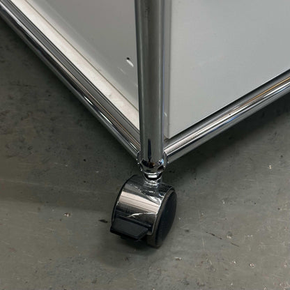 Close-up of white USM Haller Rollcontainer with chrome leg and caster wheels on gray floor