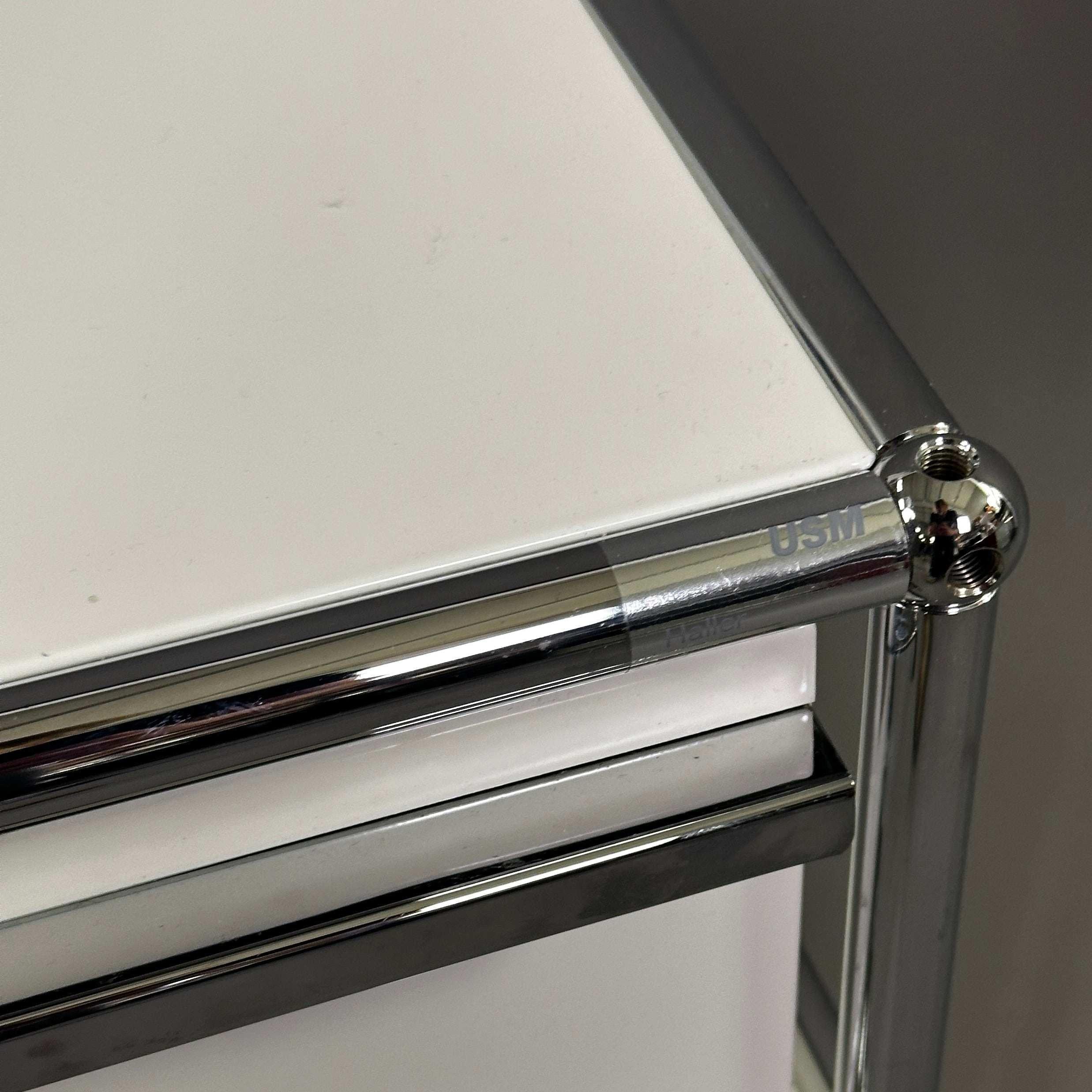 USM Haller Rollcontainer in Reinweiß mit charakteristischem Chrom-Rahmen und markantem Kugelgelenk-Design.