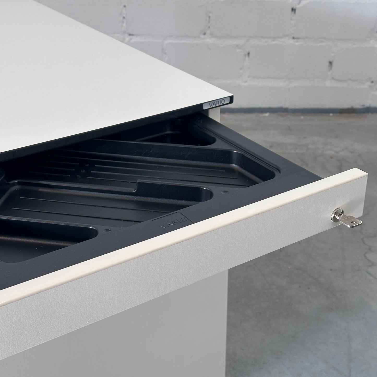 Weißer Vario Rollcontainer mit schwarzer Kante und ausgezogener Stiftschublade inklusive Schloss.