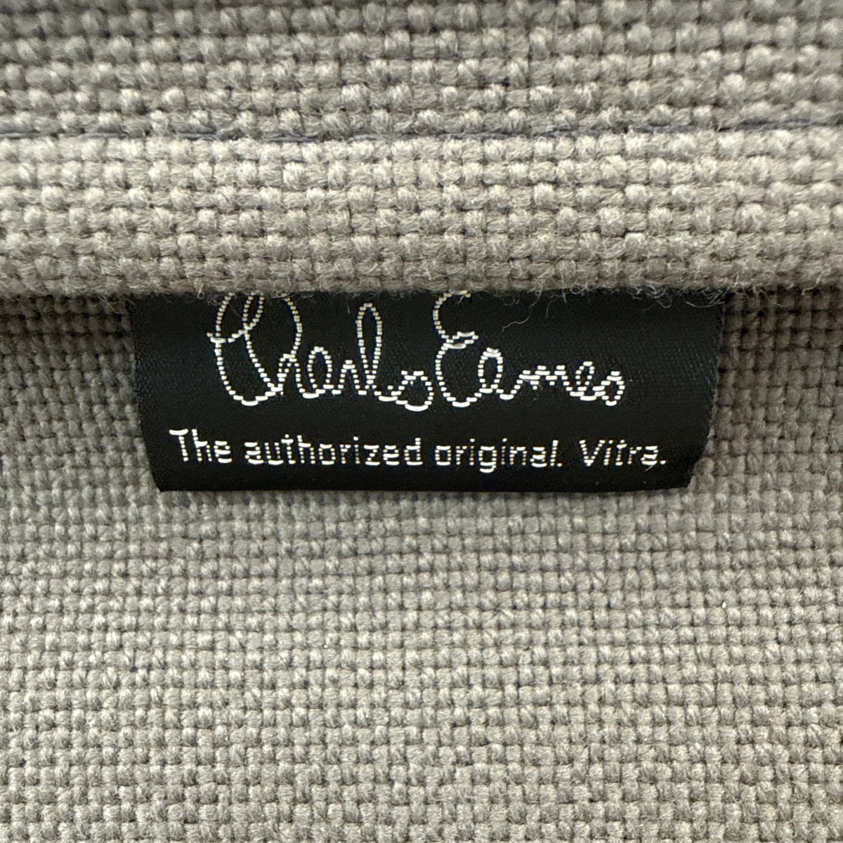 Schwarzes Charles Eames Label auf hellgrauem, strukturiertem Hopsak-Stoffbezug eines Vitra EA 119 Bürostuhls.
