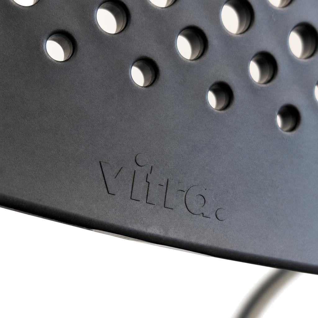 Vitra Visavis 2 Stuhl mit gelochter grauer Kunststoff-Rückenlehne und eingeprägtem Vitra-Logo.