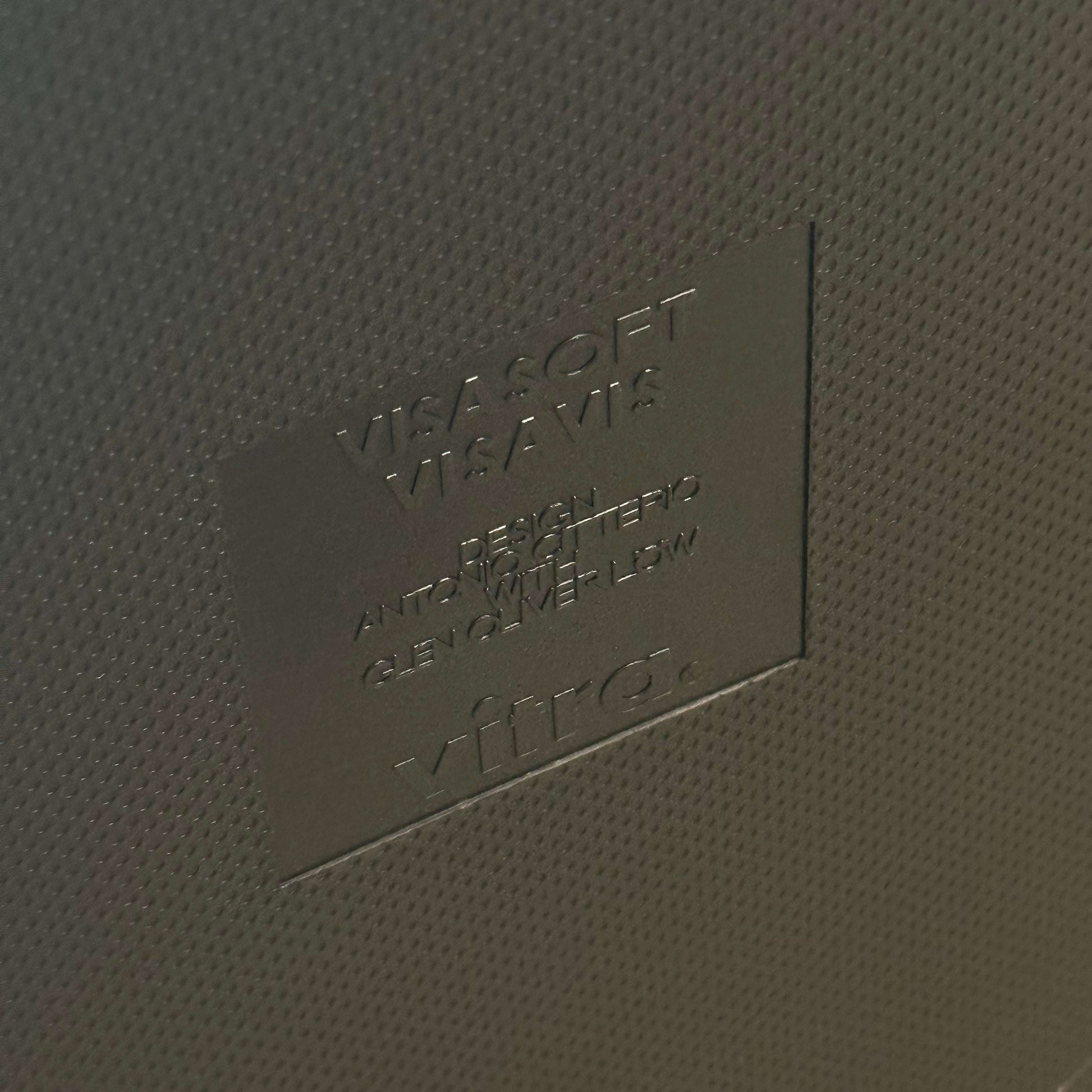 Close-up of embossed Vitra Visavis Softback logo on dark blue textured surface of chair backrest with padded upholstery.